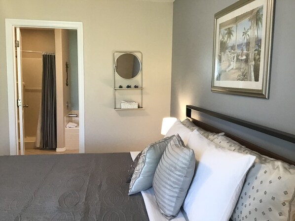 Een hotelkamer met een bed, nachtkastje, spiegel, een ingelijste foto aan de muur en een badkamer door een open deur zichtbaar.