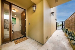 A hallway with a wooden door, a welcome mat, and a tiled floor leading to a balcony with a metal railing.