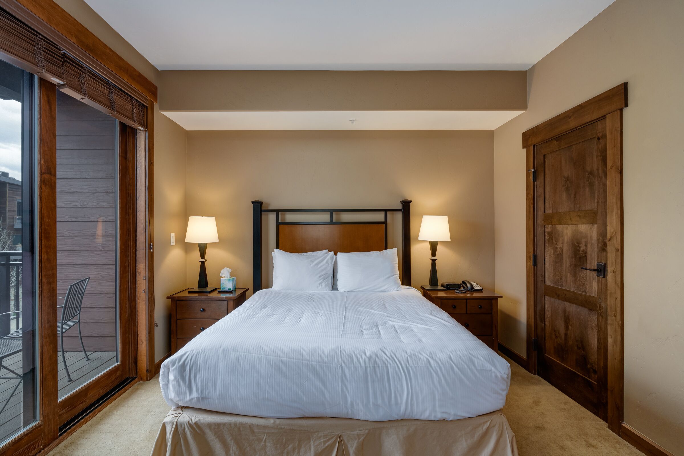 Un dormitorio con una cama grande, dos mesitas de noche con lámparas, un cabecero de madera y un ventanal corredero que da a un espacio exterior.
