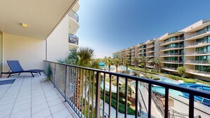 A balcony with a lounge chair, a pool area with palm trees, and a view of a multi-story hotel building. - 3br/3ba w/ Lazy River & Slide Sleeps 8 (Orange Beach)