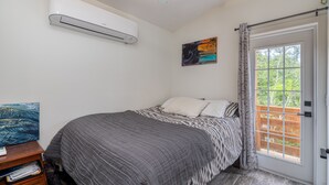 Un dormitorio con cama, aire acondicionado, un cuadro y ventana con vista a follaje.