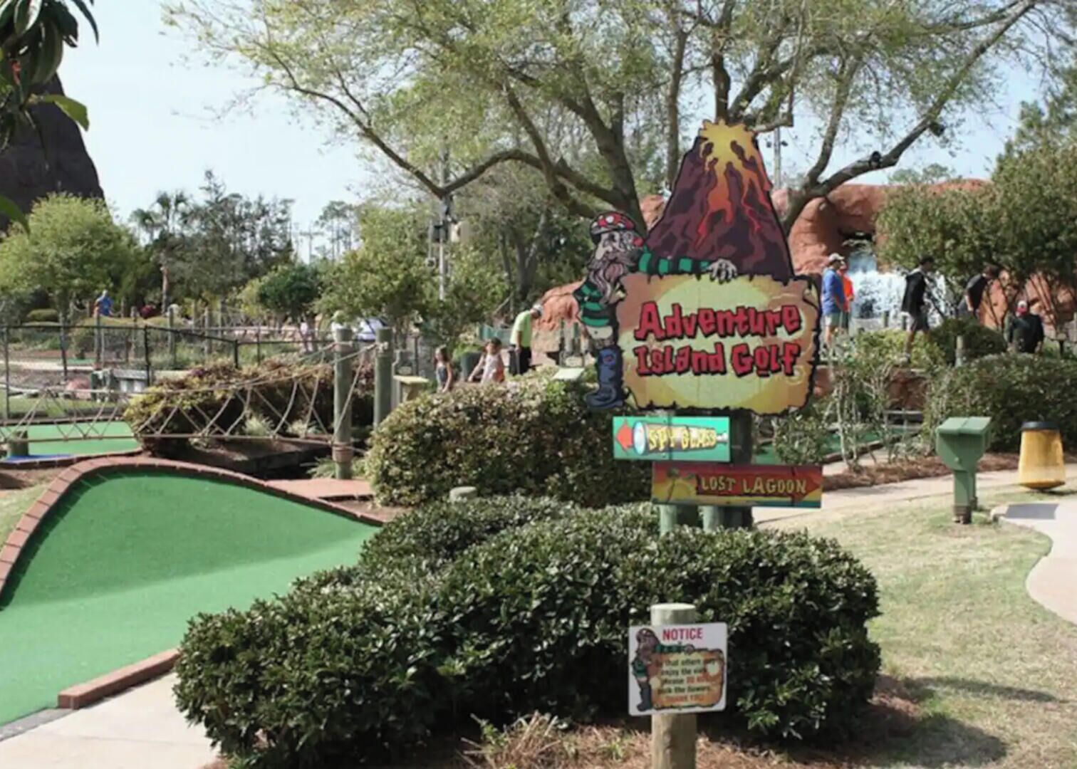 Een bord voor Adventure Island Golf met een vulkaan ontwerp, een informatiebord en een groen golfterrein.