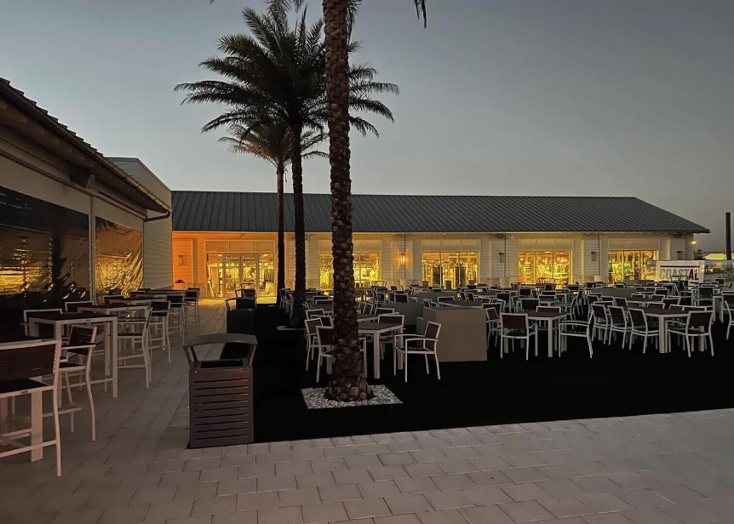 An outdoor seating area with tables and chairs, palm trees, and a building with large windows.