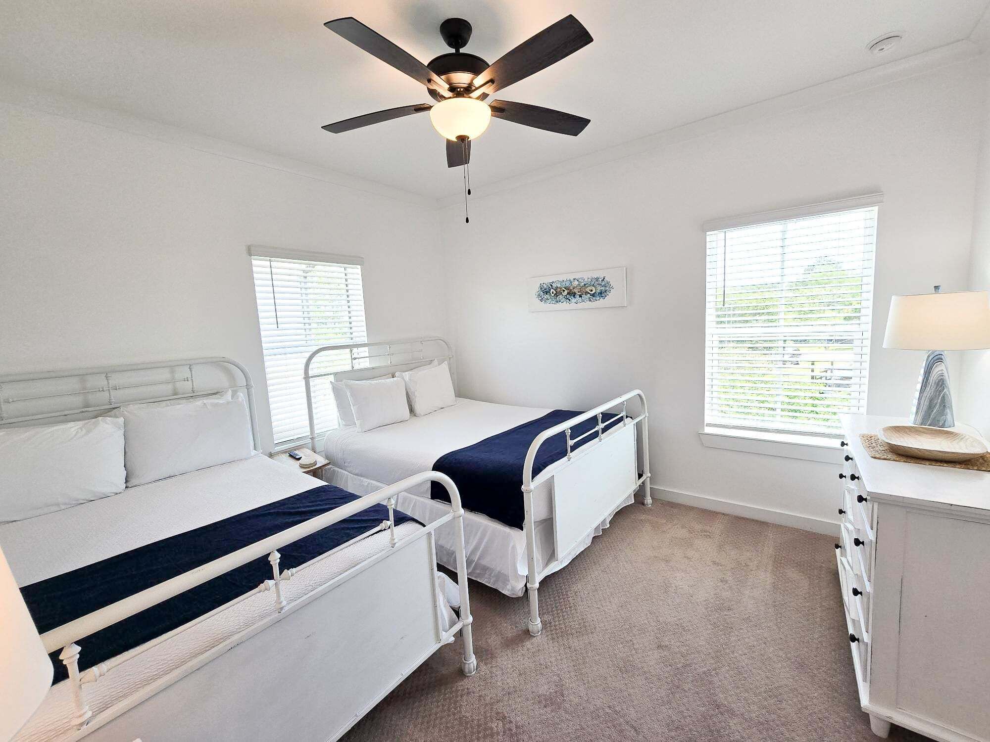 Une chambre avec deux lits, un ventilateur de plafond, une table de chevet avec une lampe et une fenêtre avec des stores.