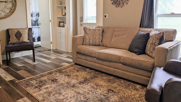 A living room with a beige sofa, patterned throw pillows, a dark brown chair, a large wall clock, and a decorative metal piece. - Farmhouse on the Prairie - You can see for miles! (valley falls)