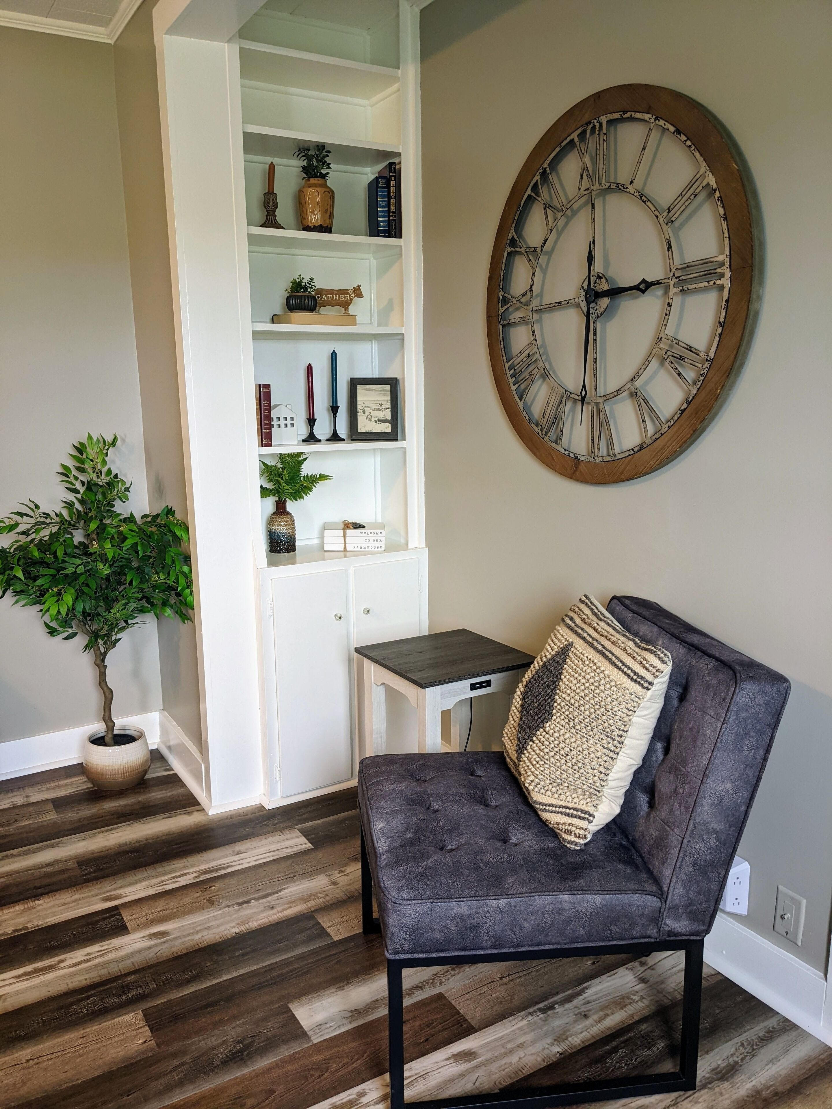 Une pièce avec un fauteuil à revêtement gris foncé, un plancher en bois, une bibliothèque blanche intégrée et une grande horloge murale.