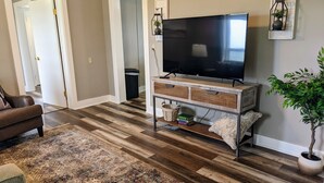 A living room with a flat-screen TV on a wooden TV stand, a patterned rug, a brown leather sofa, and a potted plant. - Farmhouse on the Prairie - You can see for miles! (valley falls)
