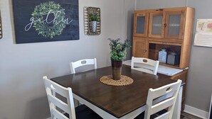 A dining area with a dark wooden table, white chairs, a woven placemat, a potted plant, and a cabinet with glass doors. - Farmhouse on the Prairie - You can see for miles! (valley falls)