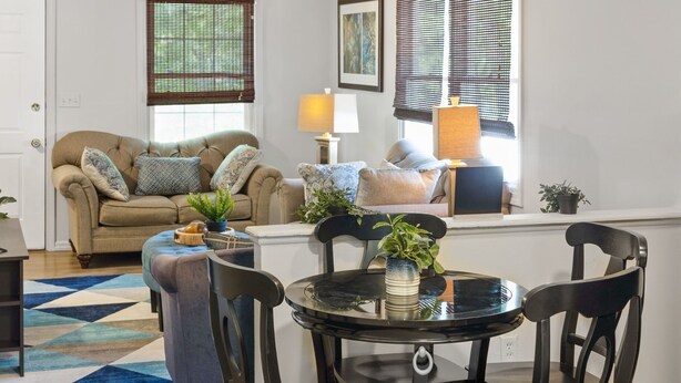 Un salon avec un canapé beige, deux fauteuils, une table basse et une table à manger avec quatre chaises. On y trouve deux lampes sur des tables d’appoint, une plante en pot sur la table basse et un tapis géométrique bleu et blanc sur le sol.