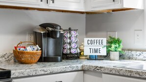 Une cuisine équipée d’une machine à café, d’un panier rempli de produits d’épicerie, d’un support à capsules de café et d’un panneau indiquant « PAUSE CAFÉ ».