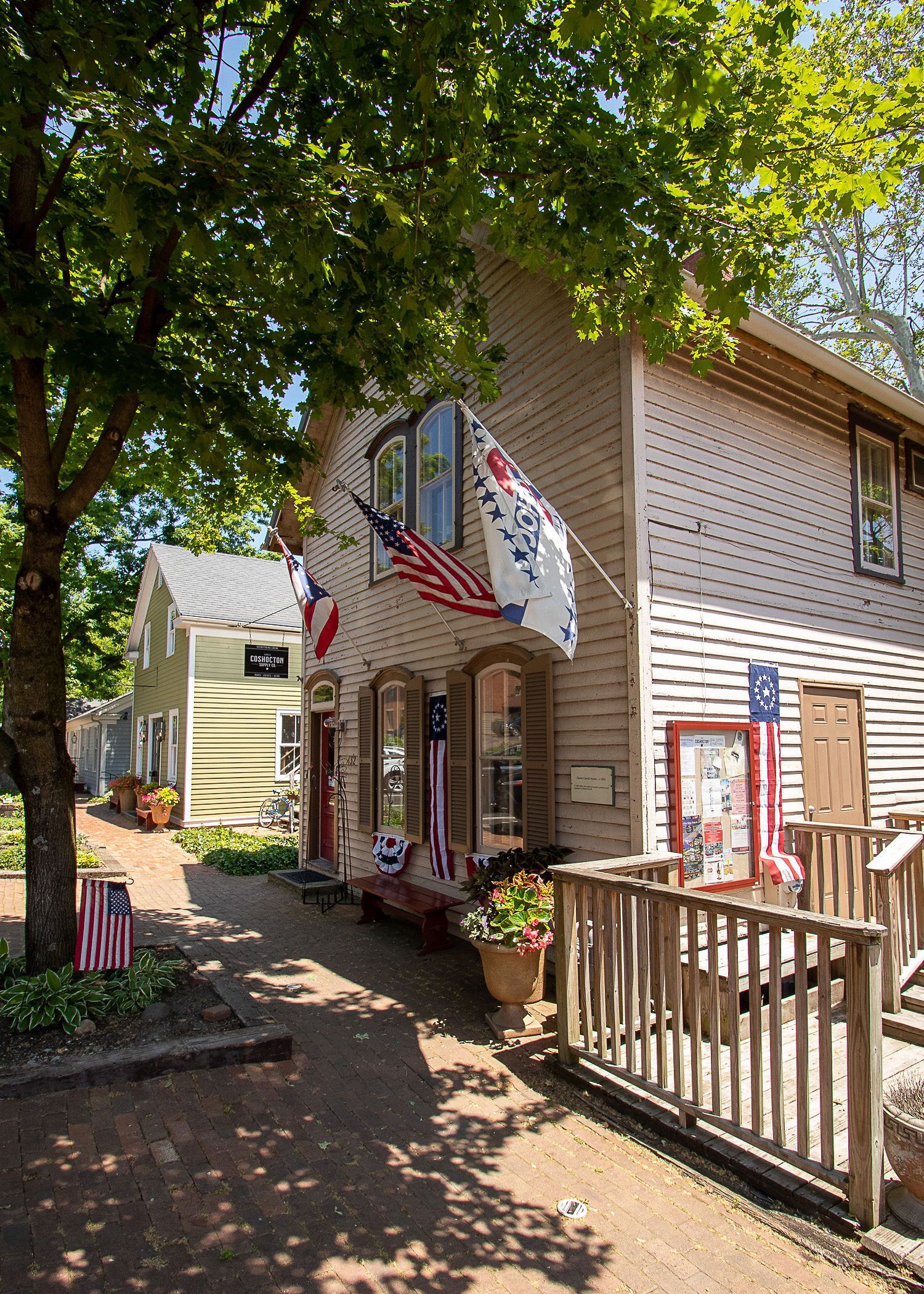 A historic building with American flags, a wooden porch, and a brick pathway.