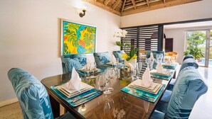 A dining area with a wooden table set for a meal, blue upholstered chairs, and a colorful tropical-themed painting on the wall. - 🏝️ Forever Summer | Gorgeous 4BDR Villa w/ Pool - Panoramic Views (Runaway Bay)