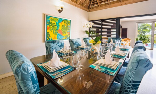 Un coin repas avec une table en bois dressée pour un repas, des chaises à l’assise rembourrée de couleur bleue et un tableau coloré sur le mur, représentant un thème tropical.