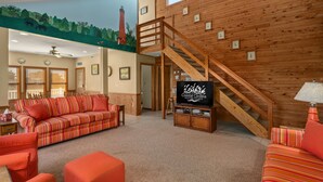 A cozy living room with a wooden staircase, a sofa, ottoman, and a television displaying 'Coastal Carolina'. - Off Crew-Oceanfront, pool, hot tub, private walkway to the beach, game room, ocean views! (Corolla)