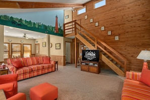 A cozy living room with a wooden staircase, a sofa, ottoman, and a television displaying 'Coastal Carolina'.