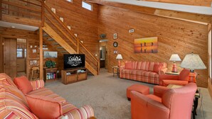 A spacious living room with a wooden interior, a staircase, a television, and a large painting on the wall. - Off Crew-Oceanfront, pool, hot tub, private walkway to the beach, game room, ocean views! (Corolla)