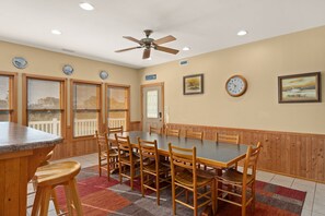 A dining area with a long table, wooden chairs, a ceiling fan, and a clock on the wall.