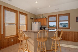 Cocina con armarios de madera, un refrigerador, un microondas y un lavamanos. Hay tres taburetes de bar de madera y una ventana con vista a una casa.
