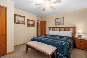 A bedroom with a bed, a bench, a nightstand, a lamp, and two paintings on the wall. - Off Crew-Oceanfront, pool, hot tub, private walkway to the beach, game room, ocean views! (Corolla)