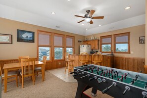 A room with a foosball table, wooden furniture, a ceiling fan, and large windows.