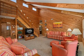 Un salón amplio con detalles de madera, una escalera, un televisor y un cuadro grande en la pared.