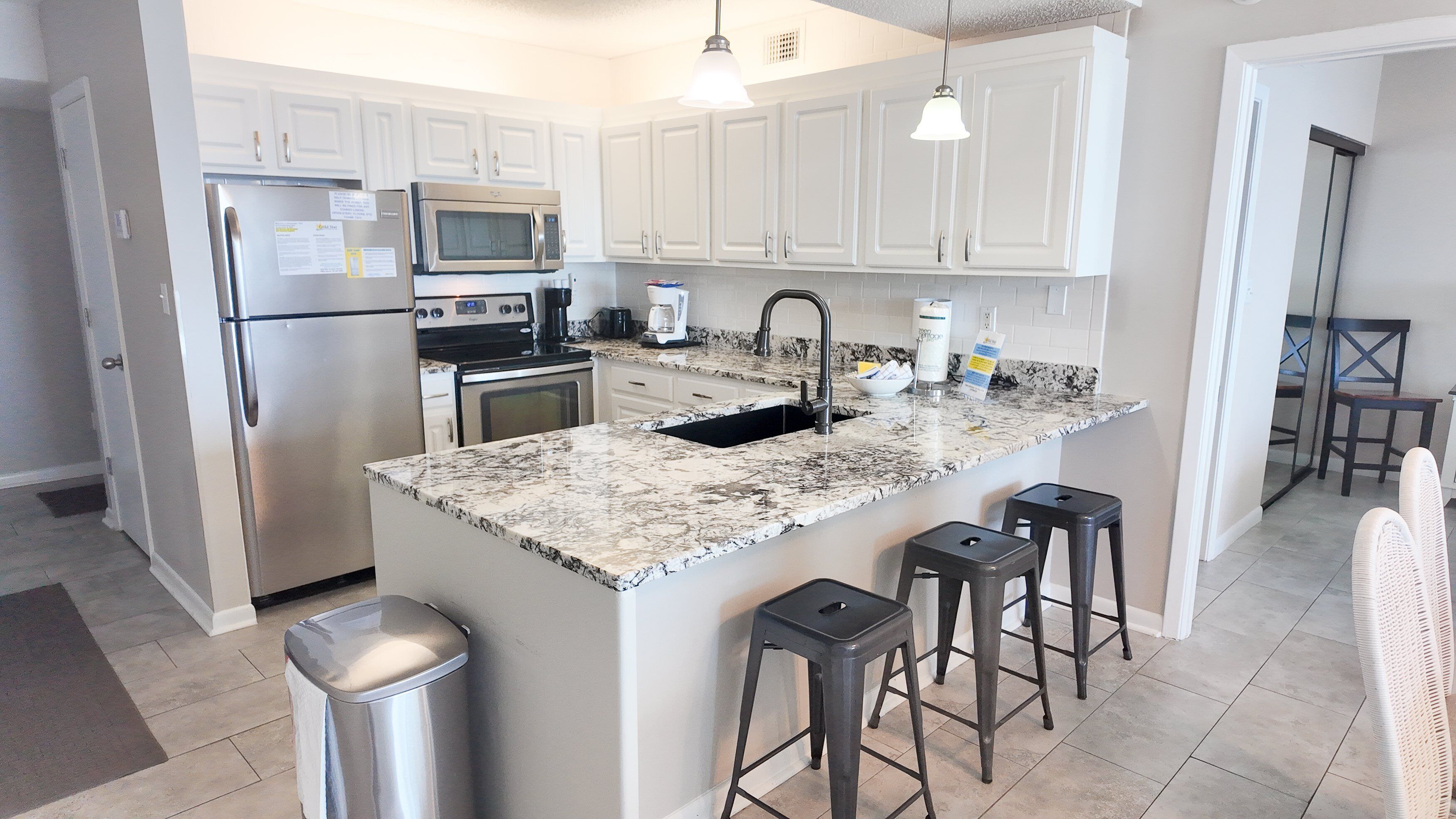 A modern kitchen with white cabinets, stainless steel appliances, a granite countertop, and a sink. There are three stools and a trash can.