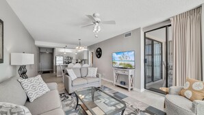 A modern living room with a sofa, coffee table, and TV, a kitchen in the background, and a large window with curtains. - Stunning Views, 3bd/2ba w/ Private Balcony (Orange Beach)