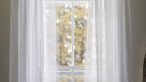 A room with a window covered by sheer curtains, a teal vase on the windowsill, and a chandelier above. - The Poplars Suite 1827: A Historic Stay (Cobourg)