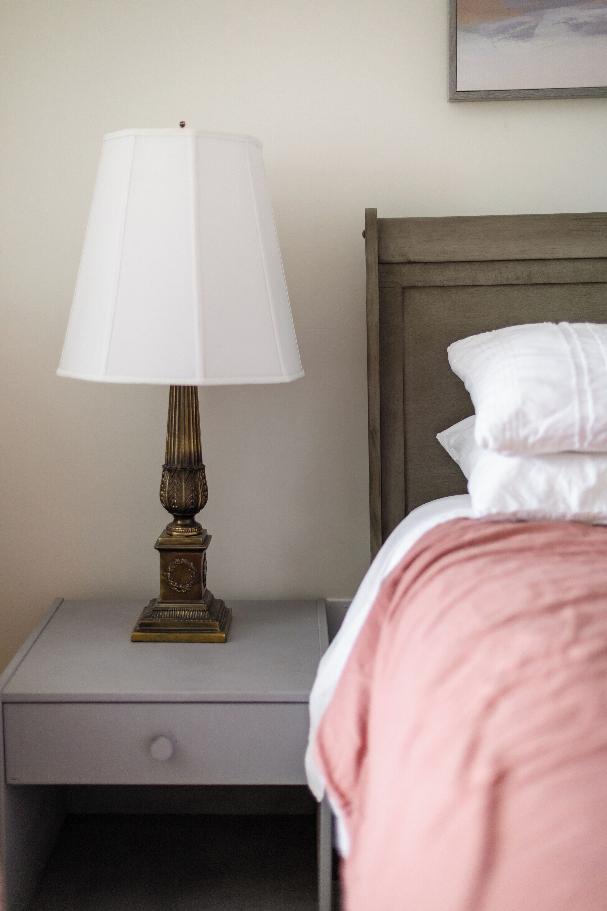 A bed with white bedding and a pink throw, a bedside lamp with a white shade, and a small drawer under the lamp.