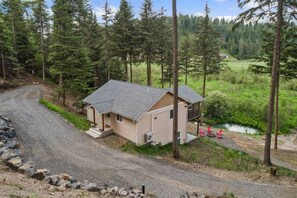 A single-story cabin with a gravel driveway, surrounded by dense forest and a small body of water.