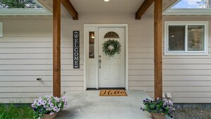 Una casa con una porta bianca, un cartello con la scritta 'WELCOME', una ghirlanda e due vasi di fiori sul portico.