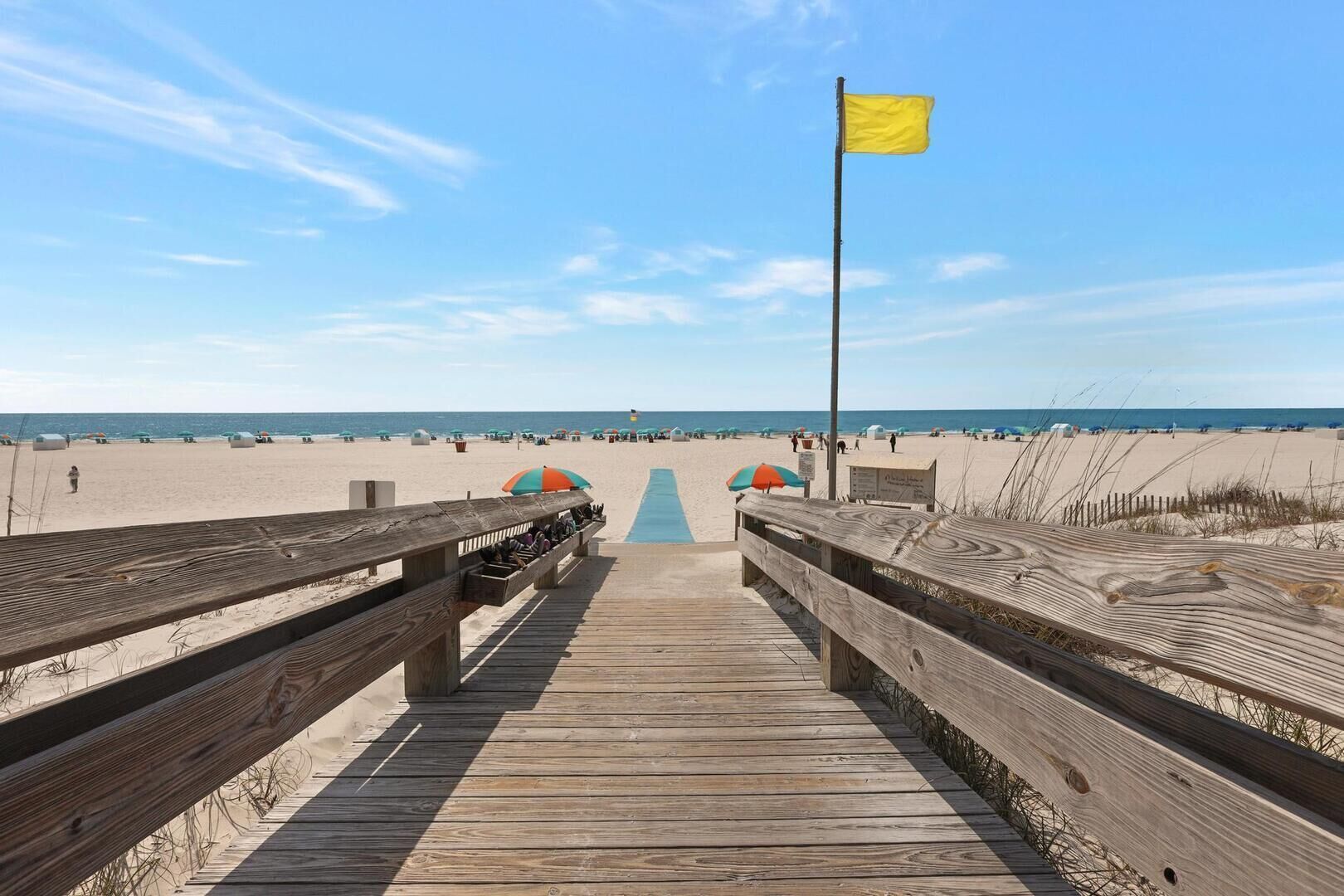 Une promenade en bois menant à une plage agrémentée de parasols et d’un drapeau.