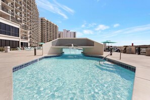A swimming pool with a water feature in the center, surrounded by a poolside area with lounge chairs and a building in the background.
