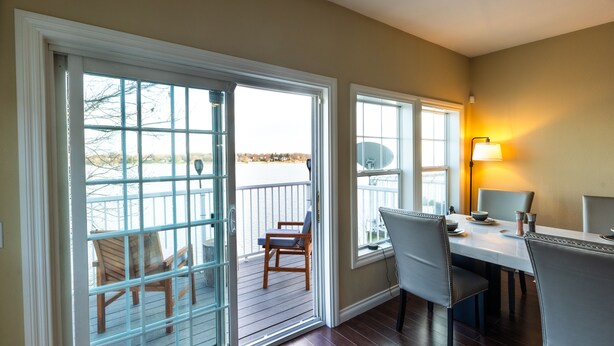 A dining area with a table and chairs, a lamp, and a view of a deck with chairs and a railing.