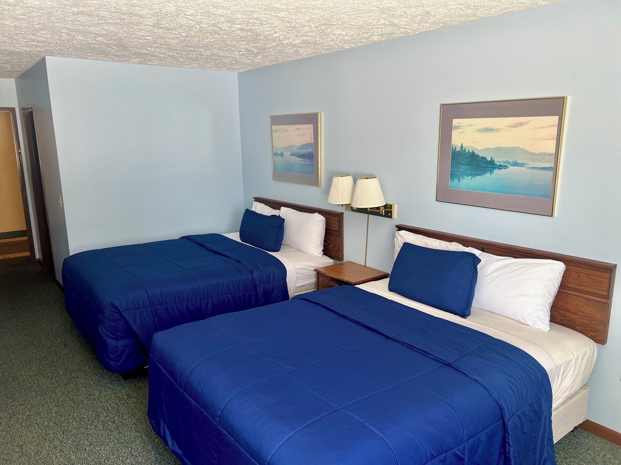 A hotel room with two beds, each with blue bedding and white pillows. There are two bedside tables with lamps, and the walls are painted light blue.
