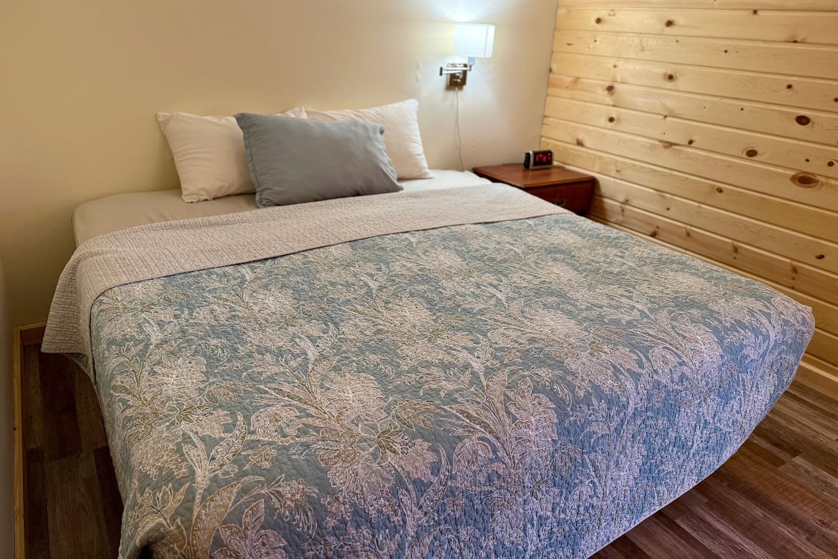 Un lit recouvert d’une couvre-lit à motifs floraux, une table de chevet avec une horloge et un mur en bois.