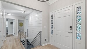 Un couloir moderne avec un plancher en bois, des murs blancs, un escalier en métal et une porte blanche ornée de panneaux de vitrail.