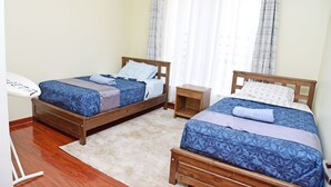 A bedroom with two wooden beds, a small bedside table, and a white chair. - Luxurious 2BR Retreat with Upscale Touches and 24/7 Security (Nairobi)
