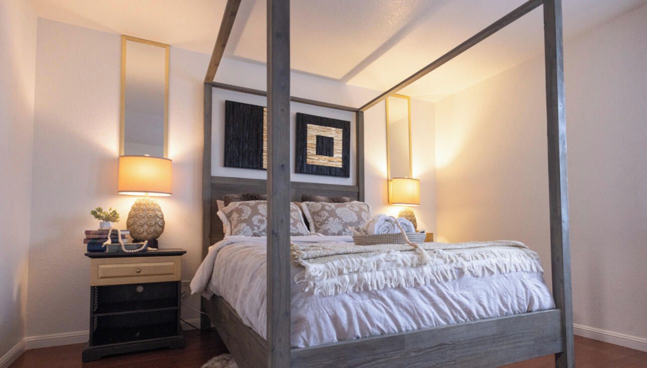 A four-poster bed with a wooden frame, a bedside table with a lamp, and a wall-mounted mirror.