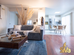 A modern living room with a grey sofa, a coffee table with books and a glass, and a dining area with a table and chairs.