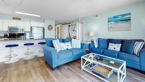 A living room with a blue sofa, a glass coffee table, and a kitchen in the background. - Beach View 3br/2ba W/pools Sleeps 8 (Orange Beach)
