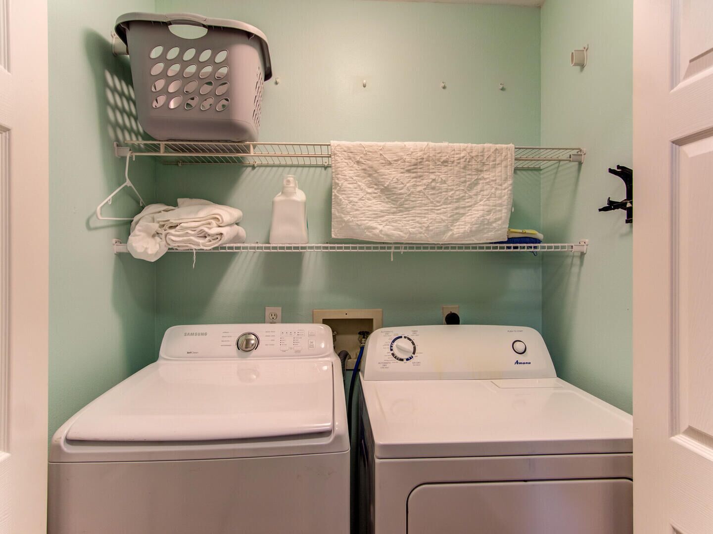 A laundry room with two white front-loading washers and dryers, a metal shelf with folded towels and a laundry basket, and a light green wall.