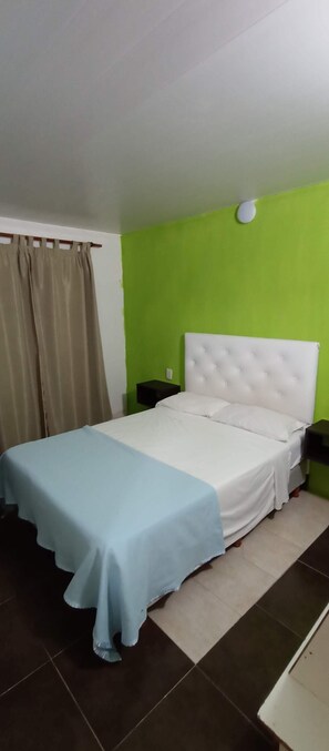 Una cama con cabecera blanca y colcha azul en una habitación con paredes verdes y suelo de baldosas.