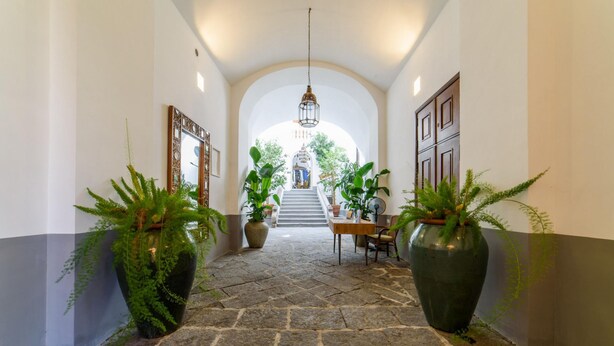 En hall med stengolv och stora krukväxter, en trappa som leder upp till ett rum med utsikt och en hängande lampa.