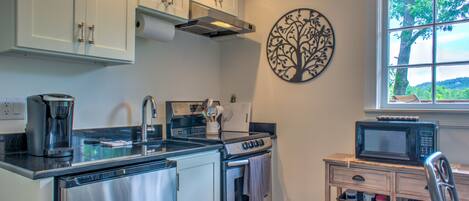 A compact kitchen with a stainless steel refrigerator, a built-in oven, a microwave, and a coffee maker. There is a window with a view of trees and a decorative wall art piece.
