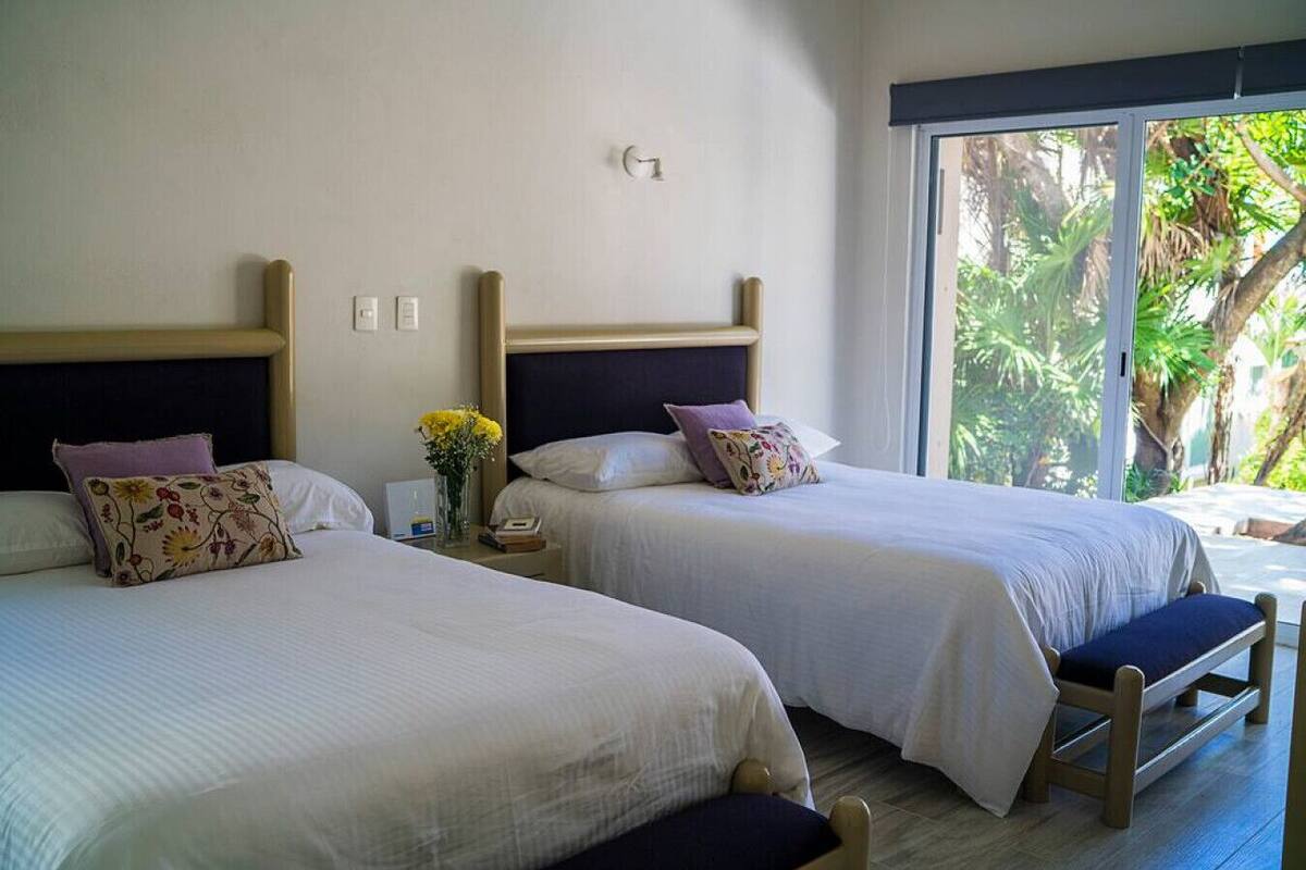 Two single beds with wooden headboards, a nightstand with a vase of flowers, and a view of greenery through a large window.