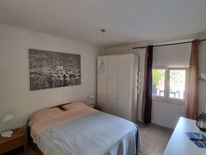 A bedroom with a bed, a wardrobe, a nightstand, a desk, a window with curtains, and a wall-mounted picture.