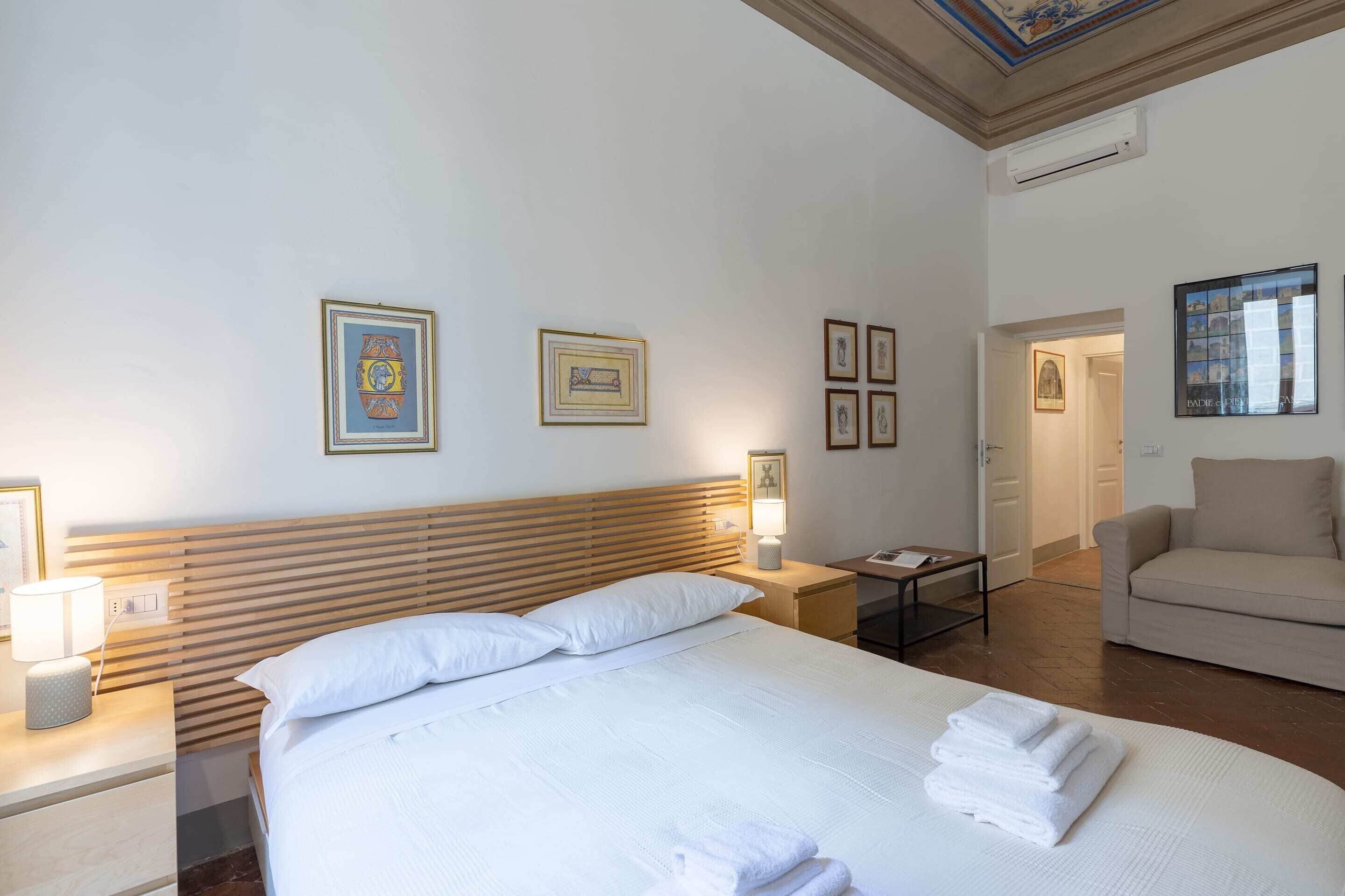 A hotel room with a bed, bedside tables, a lamp, a chair, and framed pictures on the wall.