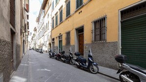 A narrow street lined with parked scooters and buildings with barred windows and doors. - Pitti Suite (Florence)