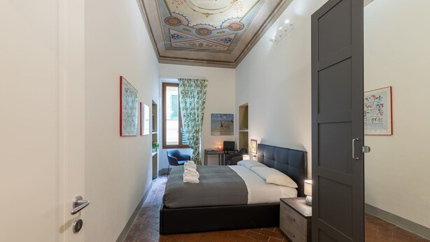 A bedroom with a large bed, a desk, and a chair. There is a door on the right side. - Pitti Suite (Florence)